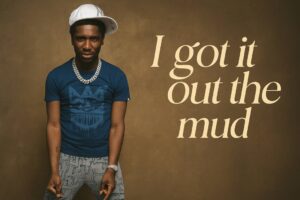 A digital photograph features PGF Nuk standing confidently indoors, wearing a blue t-shirt, patterned jeans, and a tilted white cap, with the quote “I got it out the mud” elegantly displayed beside him